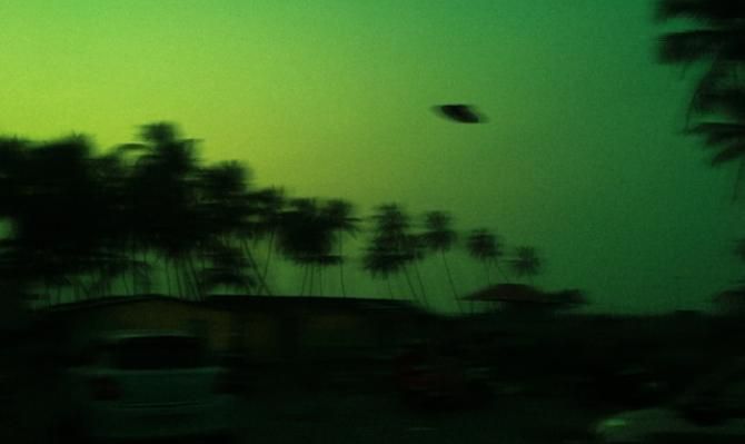 Vista nocturna de un cielo verde, con palmeras recortadas sobre el fondo, en el que se divisa un platillo volador. Photo by Albert Antony on Unsplash