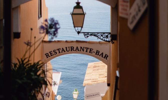 Callejuela de un pueblo marítimo con el mar de fondo y con carteles de "restaurante" para ilustrar el análisis económico de CaixaBank Research sobre el sector turístico y la restauración en España